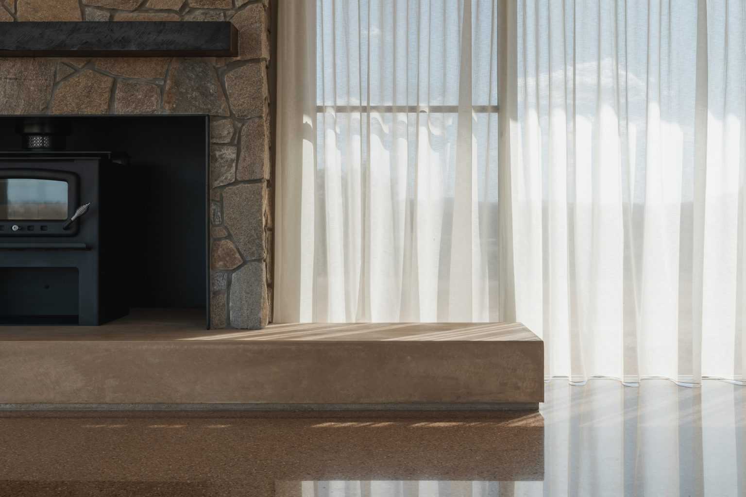 An internal frame of Table Top House featuring a stacked stone fireplace and sheer curtains